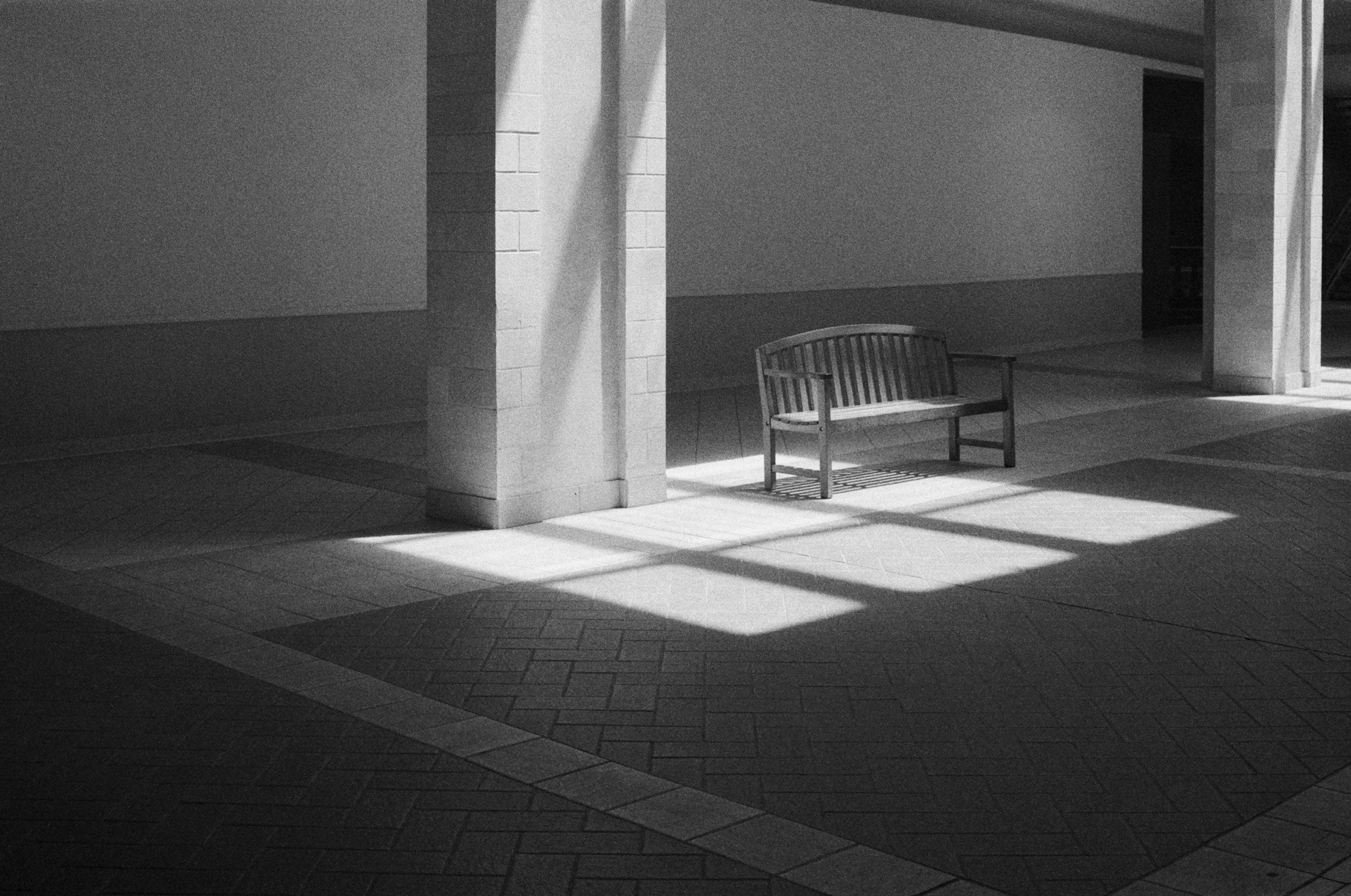 Monochrome architectural light and shadow with a solitary wooden bench for Toffee Quiet Luxury mood