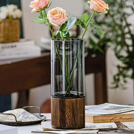 Clear vase with wooden base holding pink roses on a table.