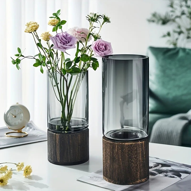 Two glass vases with wooden bases on a table, one containing flowers.